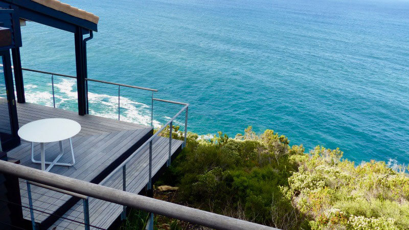 A picture of the spectacular view from our Bee's Knees Garden Route luxury villa accommodation, where you can see the ocean's waves breaking below the cliff top, and our beautiful green vegetation